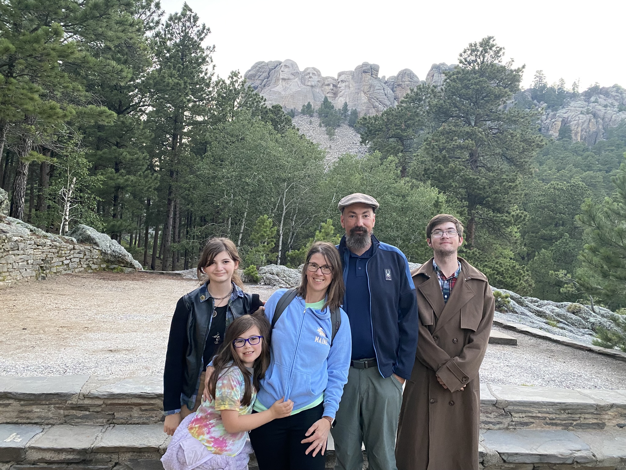 Jason Osborne and his family pose for a photo in front of Mount Rushmore National Memorial. From left to right: his teenage daughter wearing a black leather jacket, his younger daughter (with blue glasses) in a colorful tie-dye shirt and purple skirt sitting on a stone ledge, his wife smiling in a light blue "Maine" hoodie and carrying a backpack, Jason Osborne (bearded, wearing a flat cap and dark jacket), and his son (with glasses and a beard) in a brown trench coat. The iconic faces of presidents Washington, Jefferson, Roosevelt, and Lincoln are carved into the mountain behind them, surrounded by dense pine forests under a cloudy sky.