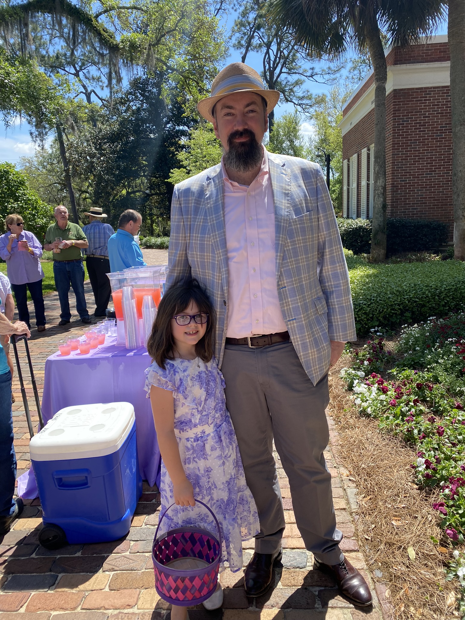 Jason Osborne, dressed in a light plaid suit jacket, pink dress shirt, gray trousers, and straw fedora, stands smiling with his young daughter at an outdoor community event, likely an Easter egg hunt. His daughter, wearing glasses and a floral purple dress, holds a woven purple and pink Easter basket while grinning at the camera. In the background, a refreshment table with purple tablecloth, colorful drinks, and a cooler is visible, along with other attendees mingling under trees draped with Spanish moss on a sunny day.