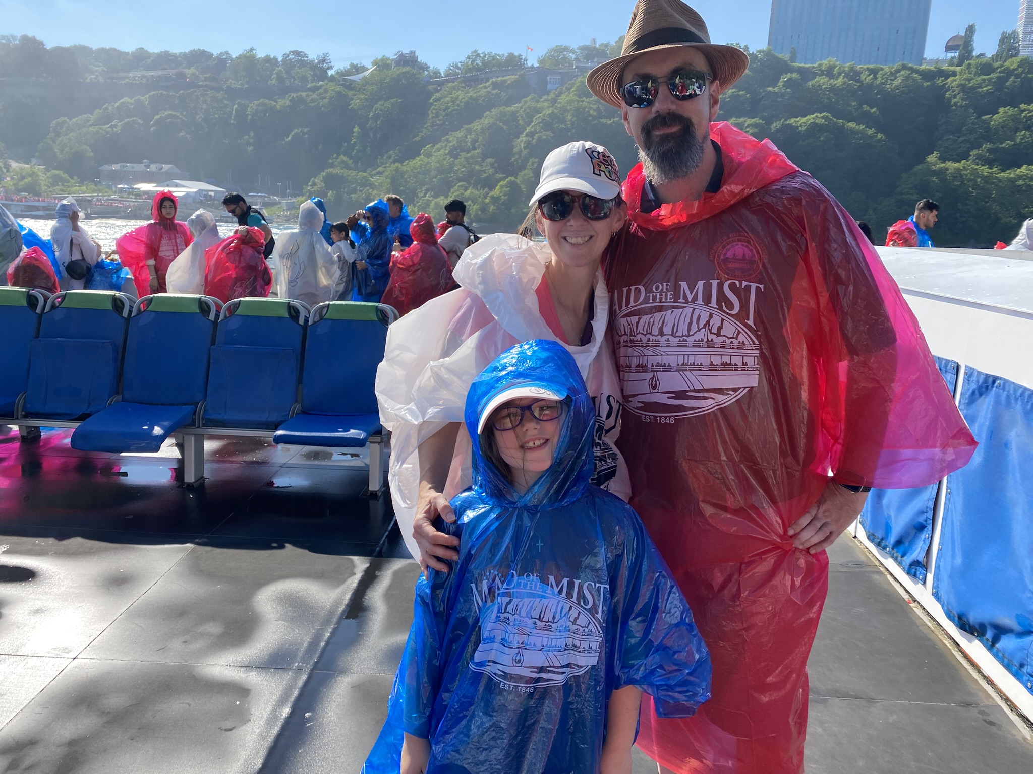 Jason Osborne, his wife, and one of their daughters pose smiling on the deck of a Maid of the Mist boat tour at Niagara Falls. All three are wearing colorful plastic rain ponchos for protection from the spray—the daughter in blue, the wife in clear/white, and Jason in red—along with sunglasses and hats (Jason in a straw fedora). The misty falls are visible in the background on the left, with lush green shoreline and other poncho-clad tourists seated on blue benches behind them under a bright, sunny sky.